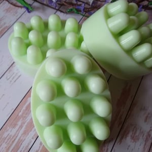 以下が含まれることがあります： 緑色のマッサージ石鹸が3つ、片側に盛り上がった突起があります。石鹸は木製の表面に半円形に配置されています。