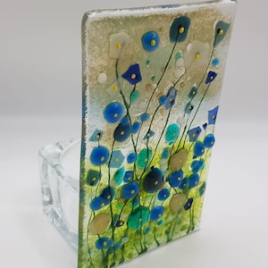 May include: A glass tealight holder with a blue, green, and yellow floral design. The holder is made of clear glass and has a square shape.