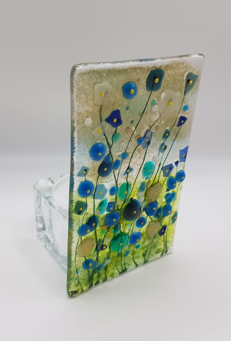 May include: A glass tealight holder with a blue, green, and yellow floral design. The holder is made of clear glass and has a square shape.