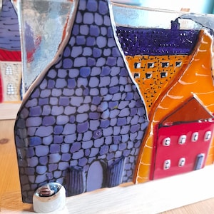 May include: A stained glass artwork featuring a row of colourful houses with purple, orange and red accents. The houses are made of glass and have a textured, mosaic-like appearance. The piece is mounted on a wooden base.