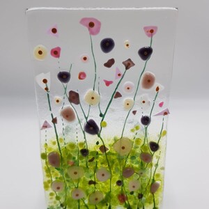 May include: A glass art piece depicting a field of wildflowers. The flowers are pink, white, and purple, with green stems and leaves. The background is a light blue.