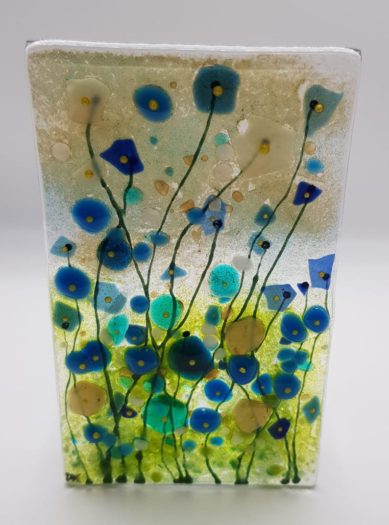 May include: A glass art piece featuring a field of blue, green, and yellow flowers. The flowers are abstract and have a textured look. The background is a light beige color.