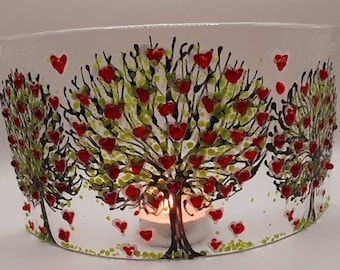 May include: A white glass candle holder with a tree design. The trees have black branches, green leaves, and red heart-shaped fruit. A lit candle is inside, casting a warm glow. The design is repeated around the holder.
