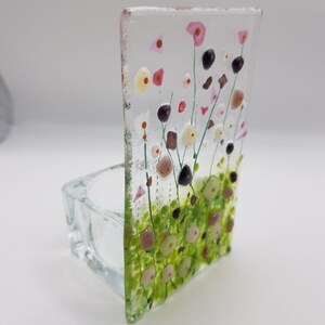 May include: A clear glass candle holder with a floral design. The design features pink, white, and green flowers with black centers. The holder is square and has a small opening at the top for a candle.