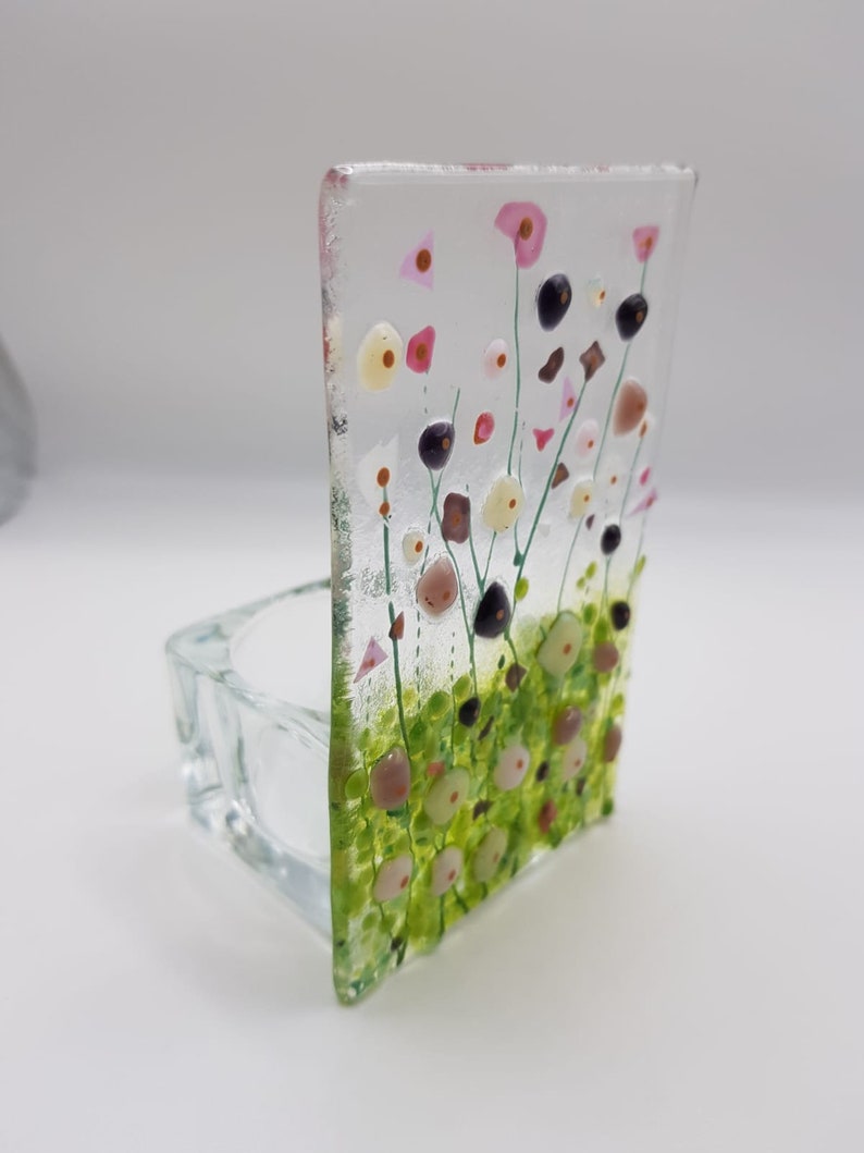 May include: A clear glass candle holder with a floral design. The design features pink, white, and green flowers with black centers. The holder is square and has a small opening at the top for a candle.