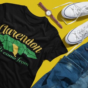 May include: Black t-shirt with the text "Clarendon Mi come from" in gold and green, featuring a map outline. Includes white sneakers, a brown leather watch, and blue jeans on a yellow background.