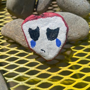 May include: A painted rock in the shape of a heart, featuring a white face with black eyes and a small black mouth. Blue teardrops are painted below the eyes, and the top of the heart is outlined in red. The rock is resting on a yellow metal surface.