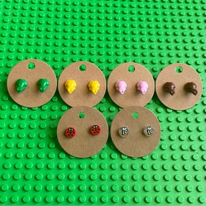 May include: Six pairs of stud earrings on brown card stock circles. The earrings feature various animal and insect designs, including turtles, ladybugs, bees, and pigs. The earrings are displayed on a green Lego baseplate.