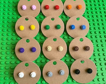 Op de afbeelding: Een verzameling kleurrijke oorstekers, elk gemaakt van een Lego-steen. De oorbellen worden gepresenteerd op kleine, ronde, bruine kartonnen kaartjes. Kleuren zijn onder meer roze, rood, oranje, geel, groen, blauw, paars, beige, bruin, wit, grijs en zwart.