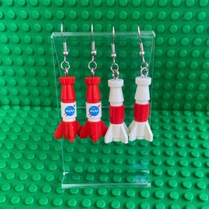 Puede incluir: Cuatro pares de pendientes novedosos con forma de cohetes rojos y blancos con el logotipo de la NASA. Los pendientes se exhiben en un soporte de acrílico transparente sobre un fondo verde.