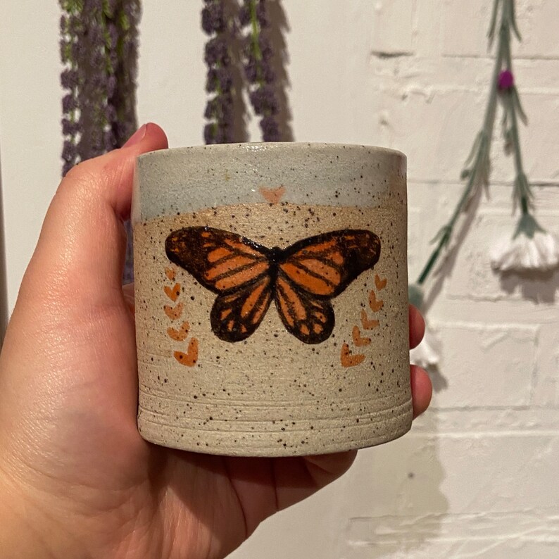 May include: A speckled ceramic mug with a butterfly design. The butterfly is orange and black, centered on the mug. The mug has a light blue top and a beige body with small brown speckles. Small orange hearts and leaves surround the butterfly.