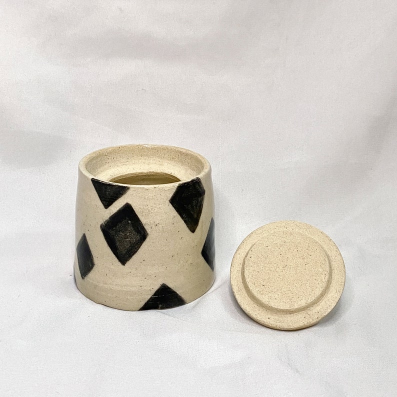 May include: A ceramic jar with a lid, featuring a cream-colored base and black diamond patterns. The jar is cylindrical with a slightly wider opening. The lid is round and flat, matching the jar's color and texture. The jar is likely handmade.