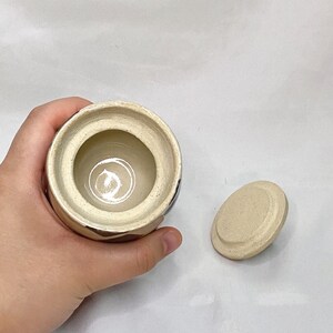 May include: A small, round ceramic jar with a lid. The jar is beige with brown geometric designs and a cream-colored interior. The lid is oval-shaped and matches the jar's color. The jar is held in a hand.