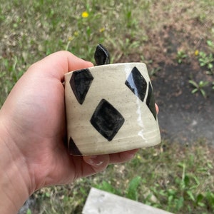 May include: A handmade ceramic jar with a lid, featuring a cream-colored base adorned with black diamond shapes. The jar is cylindrical and held in a hand, showcasing its unique, artistic design. The lid is a dark, abstract shape.