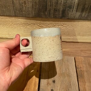 May include: A speckled ceramic mug with a square handle featuring a circular cutout. The mug has a light beige body with dark speckles and a light gray rim. The mug is held against a wooden background.