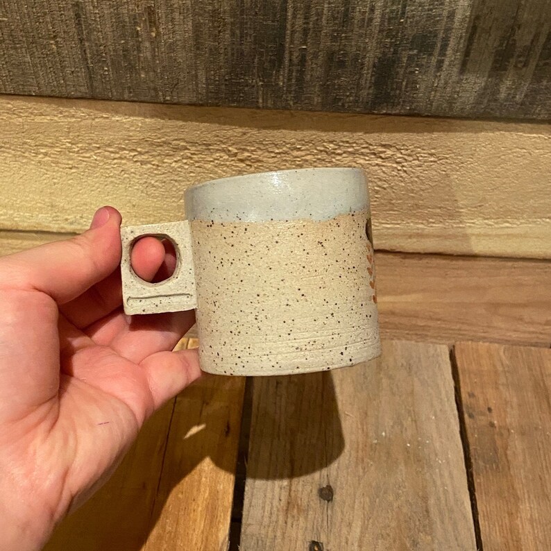 May include: A speckled ceramic mug with a square handle featuring a circular cutout. The mug has a light beige body with dark speckles and a light gray rim. The mug is held against a wooden background.