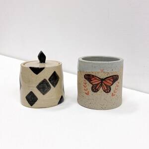 May include: Two ceramic containers. One is a small jar with a lid and black diamond pattern. The other is a cylindrical cup with a butterfly design and leaf accents. Both are on a white surface.