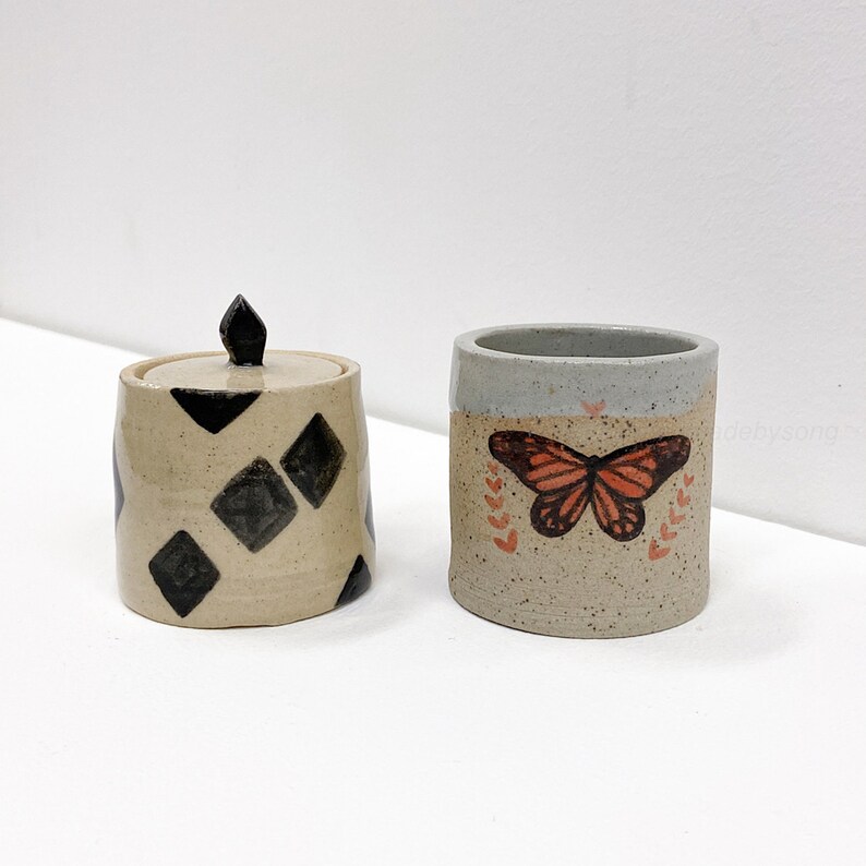 May include: Two ceramic containers. One is a small jar with a lid and black diamond pattern. The other is a cylindrical cup with a butterfly design and leaf accents. Both are on a white surface.