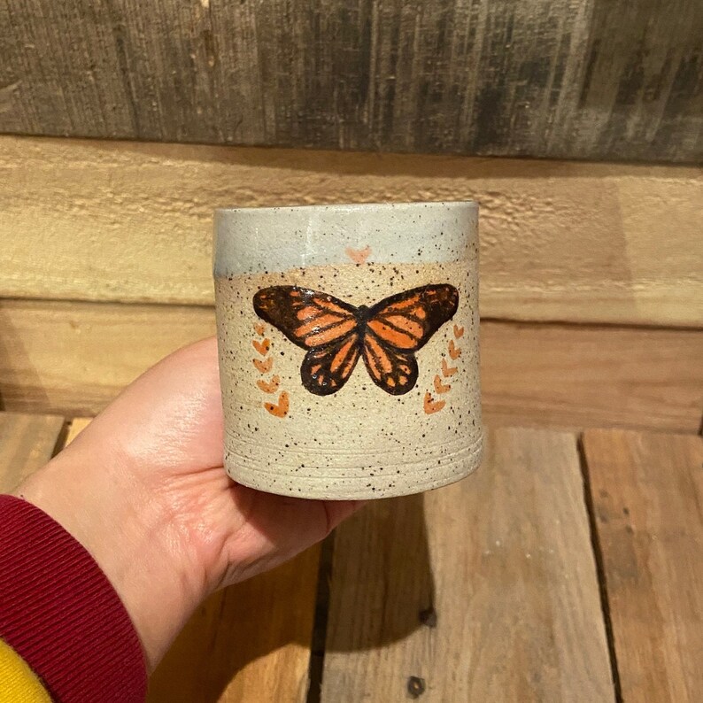 May include: A handmade ceramic mug with a butterfly design. The mug features a speckled beige body, a light blue top, and an orange and black butterfly illustration. Small orange hearts and leaf accents surround the butterfly. The mug is held against a wooden background.