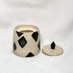 May include: A ceramic jar with a lid, featuring a cream-colored base adorned with black diamond shapes. The lid is off to the side, revealing the jar's interior. The lid has a black, pointed knob.