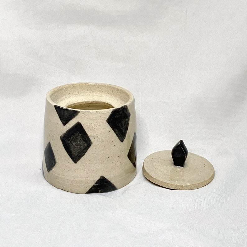 May include: A ceramic jar with a lid, featuring a cream-colored base adorned with black diamond shapes. The lid is off to the side, revealing the jar's interior. The lid has a black, pointed knob.