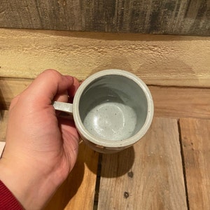 May include: A handmade ceramic mug with a light gray speckled glaze. The mug has a round body, a handle, and a textured exterior. The interior is a lighter shade of gray. The mug is designed for hot or cold beverages.