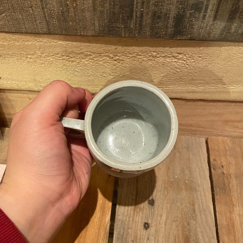 May include: A handmade ceramic mug with a light gray speckled glaze. The mug has a round body, a handle, and a textured exterior. The interior is a lighter shade of gray. The mug is designed for hot or cold beverages.