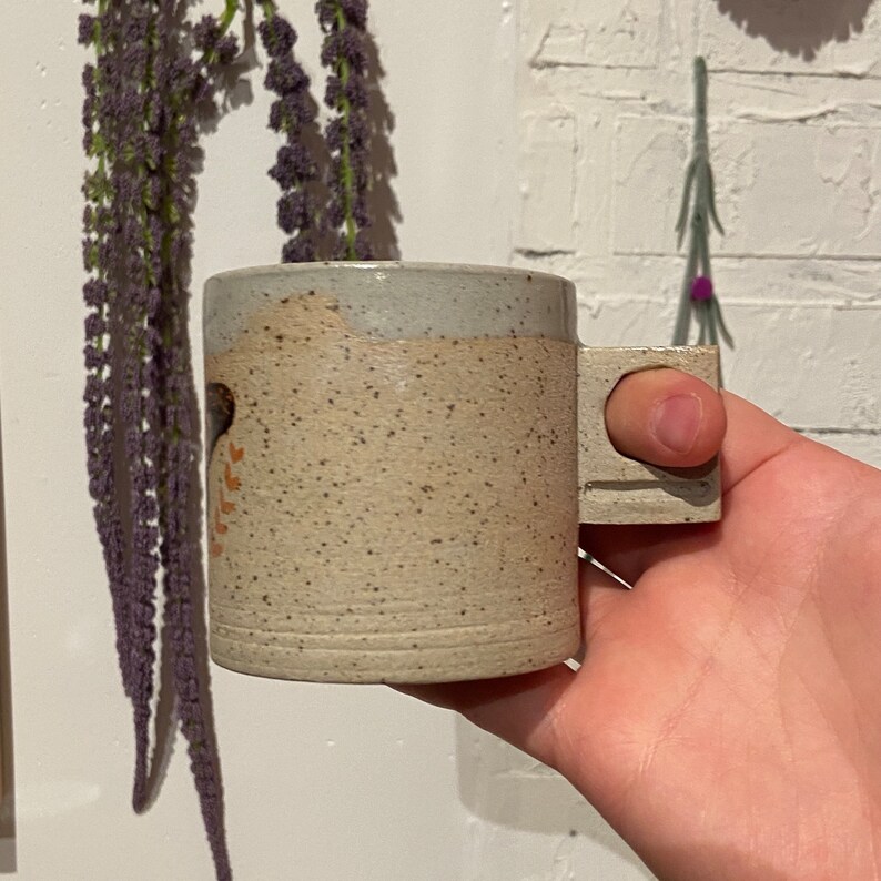 May include: A handmade ceramic mug with a speckled beige body and a light gray rim. The mug features a square handle and a simple design in orange and black. The mug is held in a hand.