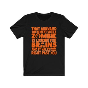 Könnte beinhalten: Schwarzes T-Shirt mit orangefarbenem Text: „THAT AWKWARD MOMENT WHEN A ZOMBIE IS LOOKING FOR BRAINS AND IT WALKS RIGHT PAST YOU“. Das Shirt hat kurze Ärmel und einen Rundhalsausschnitt.