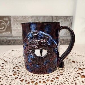 May include: A dark purple and blue ceramic mug with a textured, eye-like design. The mug has a white eye with a black pupil and a white rim.