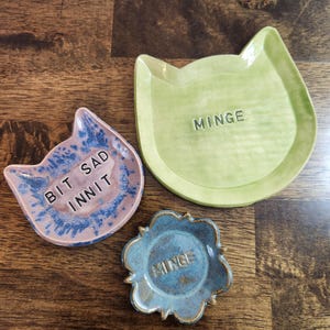 May include: Three ceramic trinket dishes in the shape of a cat's head and a flower. The cat-shaped dishes are light green and pink with the text "BIT SAD INNIT" and "MINGE". The flower-shaped dish is blue with the text "MINGE".