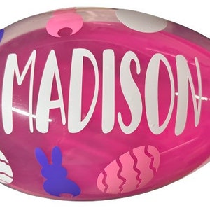 May include: Pink plastic Easter egg with a white design of bunny rabbits, eggs, and polka dots. The egg has the name "MADISON" written in white letters.