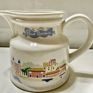 May include: A cream-colored ceramic pitcher featuring a curved handle and a pouring spout. The front displays a blue graphic with the text "la vallée de Paul Bocuse" above a colorful illustration of a village with buildings in pink, blue, and yellow.