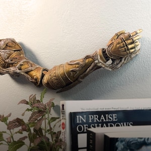 Op de afbeelding: Een gedetailleerde, goudkleurige, mechanische armsculptuur met ingewikkelde ontwerpen en een wijzende hand. De arm is op een witte muur gemonteerd, met een plant en boeken eronder. De boeken bevatten de titel "IN PRAISE OF SHADOWS".