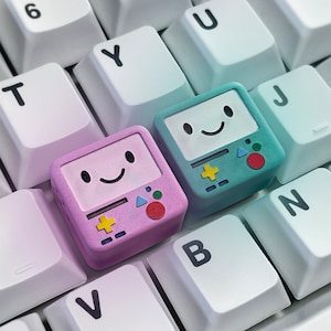 May include: Two custom keycaps with a cartoon character design, one pink and one teal, sit on a white keyboard. The keycaps have a smiling face and a game console design. The keyboard keys are white with black lettering.