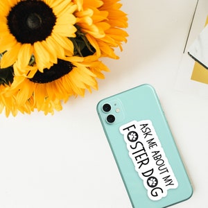 K&ouml;nnte beinhalten: Ein hellt&uuml;rkisfarbenes Smartphone mit einem wei&szlig;en Aufkleber mit dem schwarzen Text "ASK ME ABOUT MY FOSTER DOG". Im Hintergrund ein Strau&szlig; gelber Sonnenblumen.