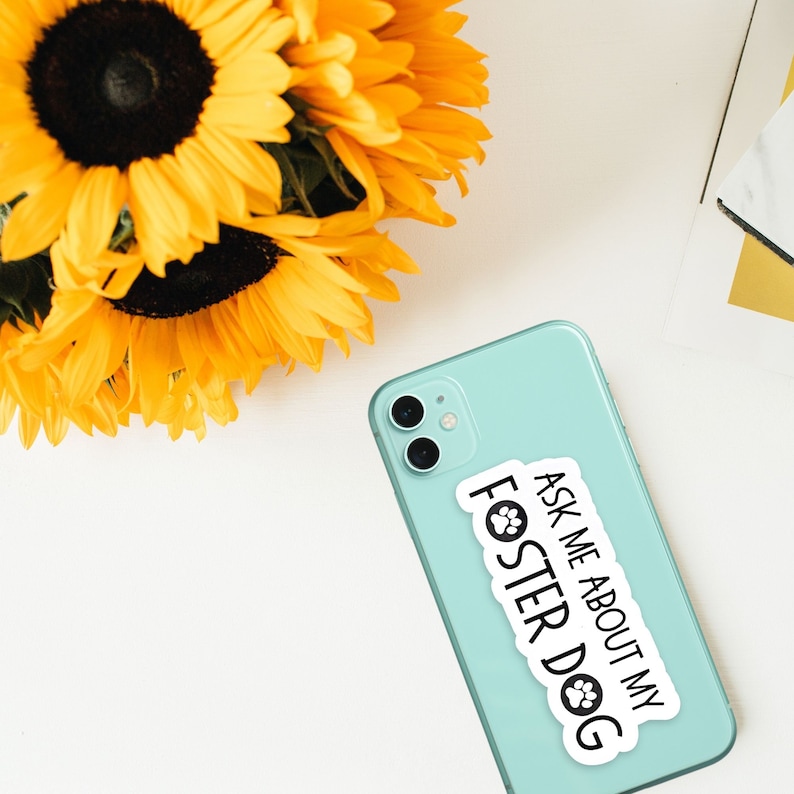 K&ouml;nnte beinhalten: Ein hellt&uuml;rkisfarbenes Smartphone mit einem wei&szlig;en Aufkleber mit dem schwarzen Text "ASK ME ABOUT MY FOSTER DOG". Im Hintergrund ein Strau&szlig; gelber Sonnenblumen.