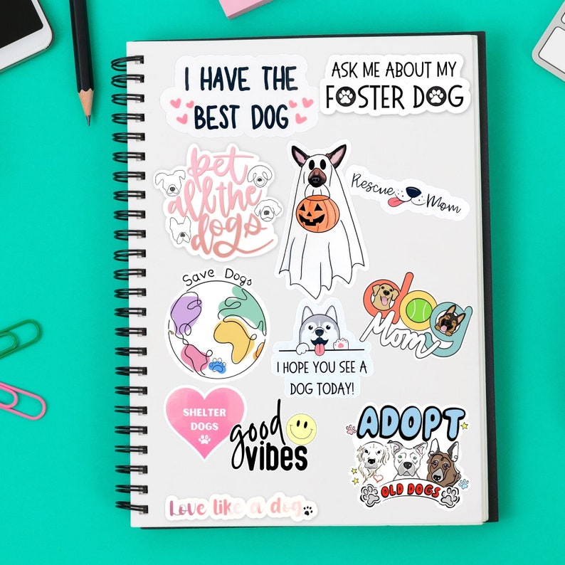 K&ouml;nnte beinhalten: Eine Sammlung von Aufklebern zum Thema Hunde auf einem Spiralnotizbuch. Die Aufkleber zeigen verschiedene Hunderassen, S&auml;tze wie "Ich habe den besten Hund" und Grafiken wie ein Geist mit einem K&uuml;rbis. Die Farbpalette umfasst Rosa, Blau und Wei&szlig;.