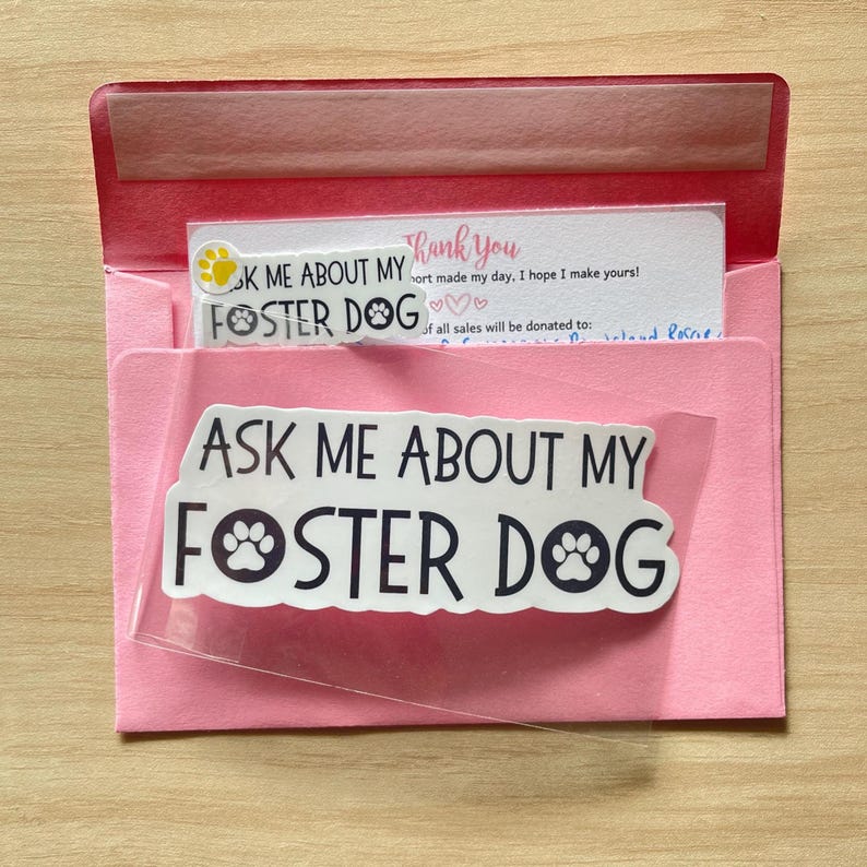 K&ouml;nnte beinhalten: Eine rosa Filzumschlag mit einem transparenten Aufkleber mit dem Text "ASK ME ABOUT MY FOSTER DOG" in schwarzer Schrift. Im Inneren befinden sich ein kleinerer Aufkleber und eine "Thank you"-Karte.
