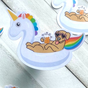 Pode incluir: Um cão amarelo usando óculos de sol em forma de coração está relaxando em um flutuador de piscina branco em forma de unicórnio com uma cauda de arco-íris. O cão está segurando um copo de cerveja.