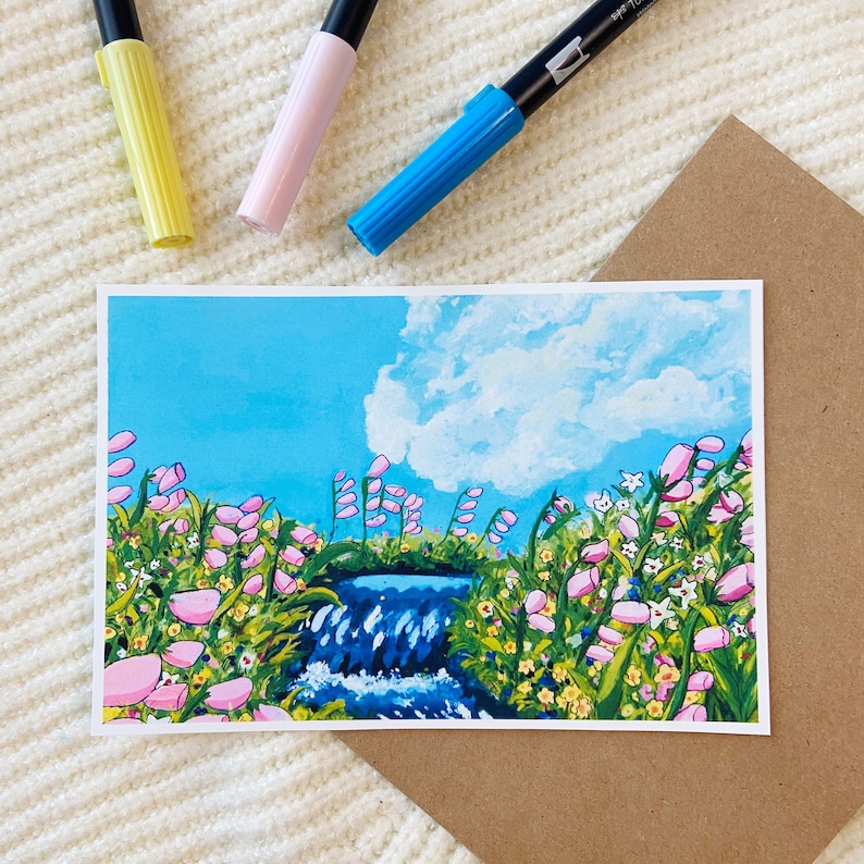 May include: A landscape art print featuring a waterfall scene with pink flowers, green foliage, and a blue sky with clouds. Three colorful markers in yellow, pink, and blue are positioned above the print. A brown envelope is also visible.