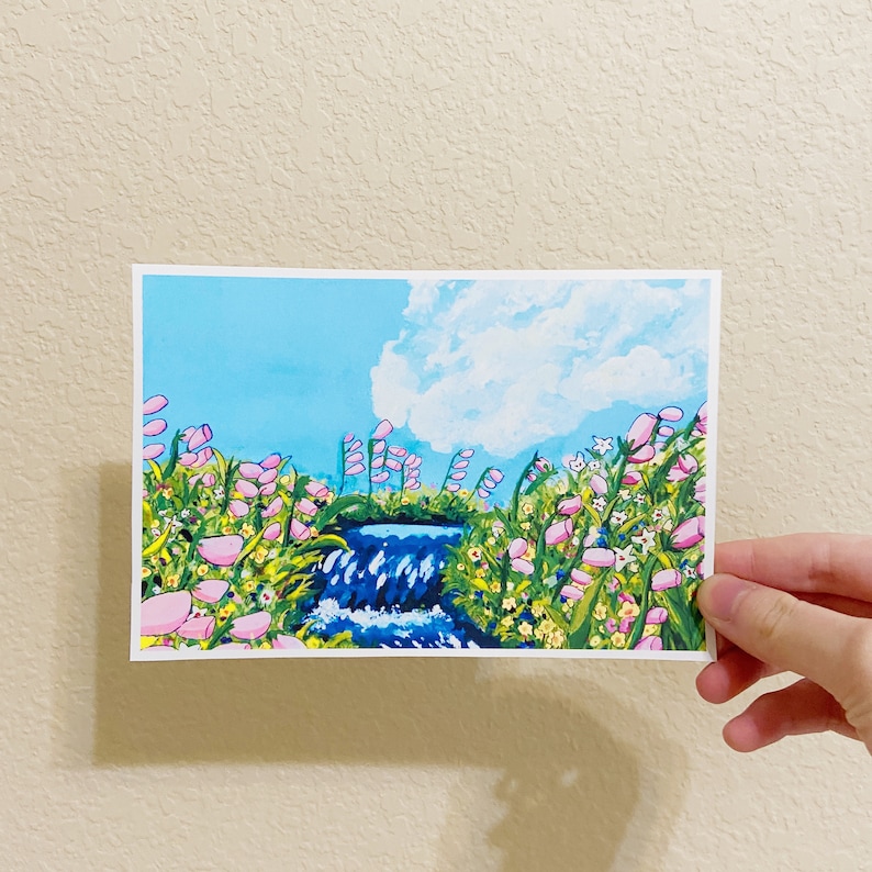 May include: A landscape painting featuring a waterfall cascading into a small stream, surrounded by pink and yellow flowers and green grass. The sky is a bright blue with a fluffy white cloud. The artwork is on a white rectangular card.