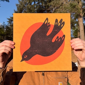 Op de afbeelding: Vierkante print met een gestileerde zwarte vogel in vlucht tegen een rode cirkel op een geel-oranje achtergrond. Het kunstwerk heeft een minimalistisch ontwerp, waarbij de silhouet van de vogel en de cirkel de belangrijkste visuele elementen vormen. De print wordt door een persoon vastgehouden.