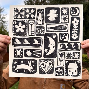 Op de afbeelding: Een zwart-wit kunstprint met blokgedrukte afbeeldingen van een konijn, maan, sterren, bloemen, een sleutel en een vlinder. De print wordt vastgehouden door een persoon die een bruine jas draagt. Het kunstwerk heeft een volkskunststijl.