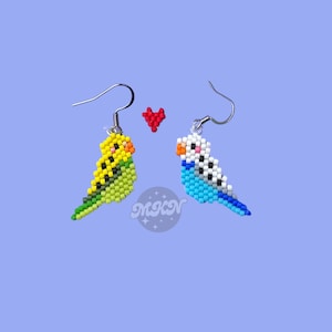 May include: A pair of beaded earrings featuring a yellow and green parakeet and a blue and white parakeet. A small red heart is between the two birds.