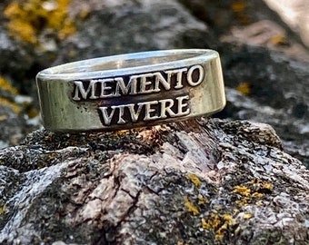 May include: A silver-toned metal ring featuring the engraved text MEMENTO VIVERE in capital letters. The band has a smooth, polished finish with dark, recessed lettering that stands out against the metallic surface of the ring.