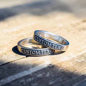May include: Two silver rings with the words "CUSTOM TEXT" engraved on the outside. The rings are stacked on top of each other, with one slightly angled. The rings are on a wooden surface.