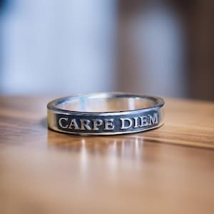 May include: A silver ring with the inscription "CARPE DIEM" engraved on the band. The ring is set against a blurred background, highlighting the inscription. The ring is a piece of jewelry.