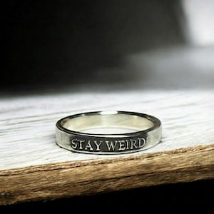 May include: A silver ring with the words "STAY WEIRD" engraved on the band. The ring is displayed on a wooden surface. The image also includes the text: "NORMAL IS BORING. EMBRACE WHAT MAKES YOU DIFFERENT."