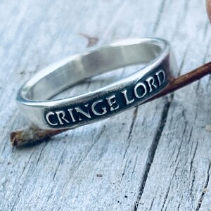 May include: A silver ring with the words "CRINGE LORD" engraved in a black font. The ring is set against a weathered, light gray wooden surface, with a small twig resting on the wood.